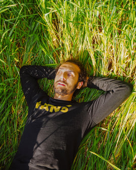 Man with brown hair and beard in long sleeve black shirt laying blissfully in bright green grass with his eyes closed.